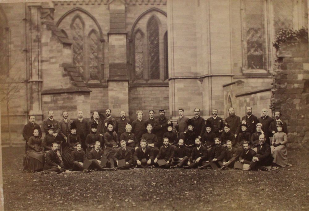 The former members of the choir and some of their wives in three rows. The wives in the centre row. Everyone is smartly dressed in black or grey suits. The women have bonnets or hats. Some of the men hold mortarboard hats in their hands. They are on the law outside the south side of the cathedral  next to the ruins of the medieval Guesten building. This is on the right of the photo. In the background is the exterior wall of the Dean's chapel and the south quire aisle.