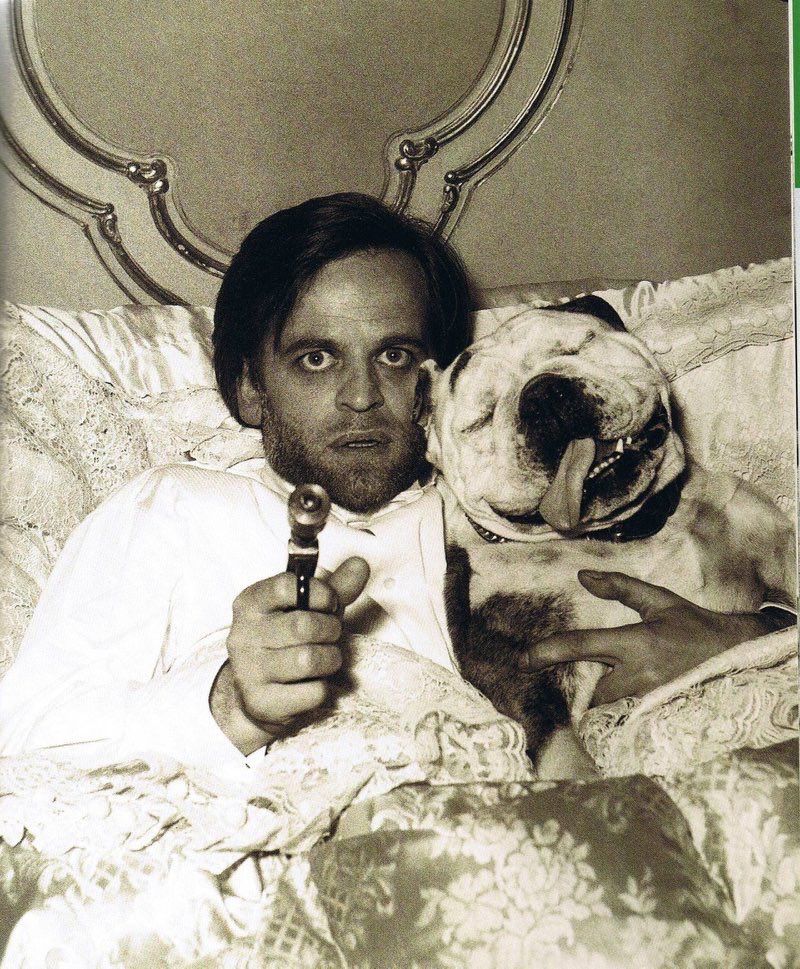 Klaus Kinski in bed with a sleeping bulldog pointing a gun at the camera