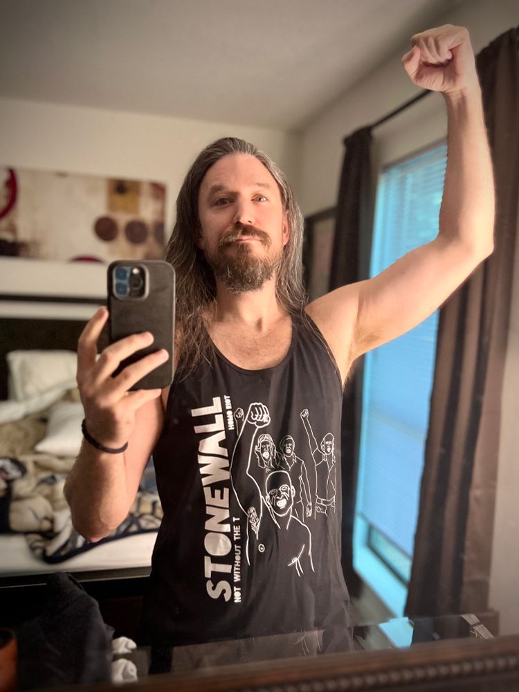 Me taking a picture of myself in the bedroom mirror, wearing a black tank that says “Stonewall, Homo Riot, Not Without The ‘T.’” The “T” in this case meaning “trans.”
