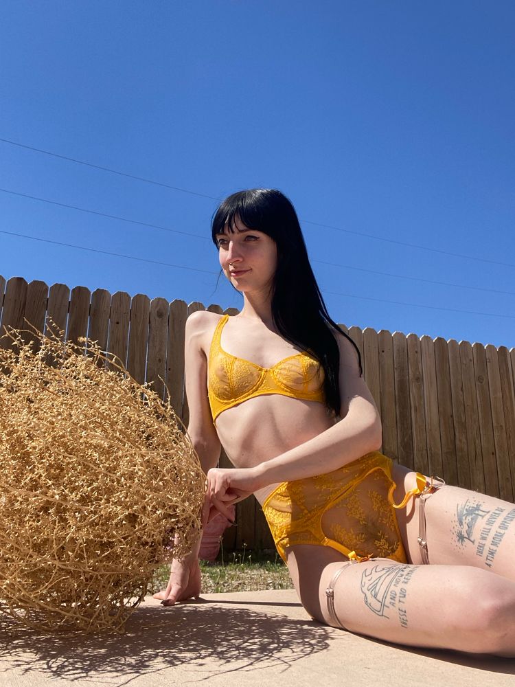 Audrey sits down outside on the ground and playfully touches a tumbleweed. She is dressed in bright yellow lingerie. 