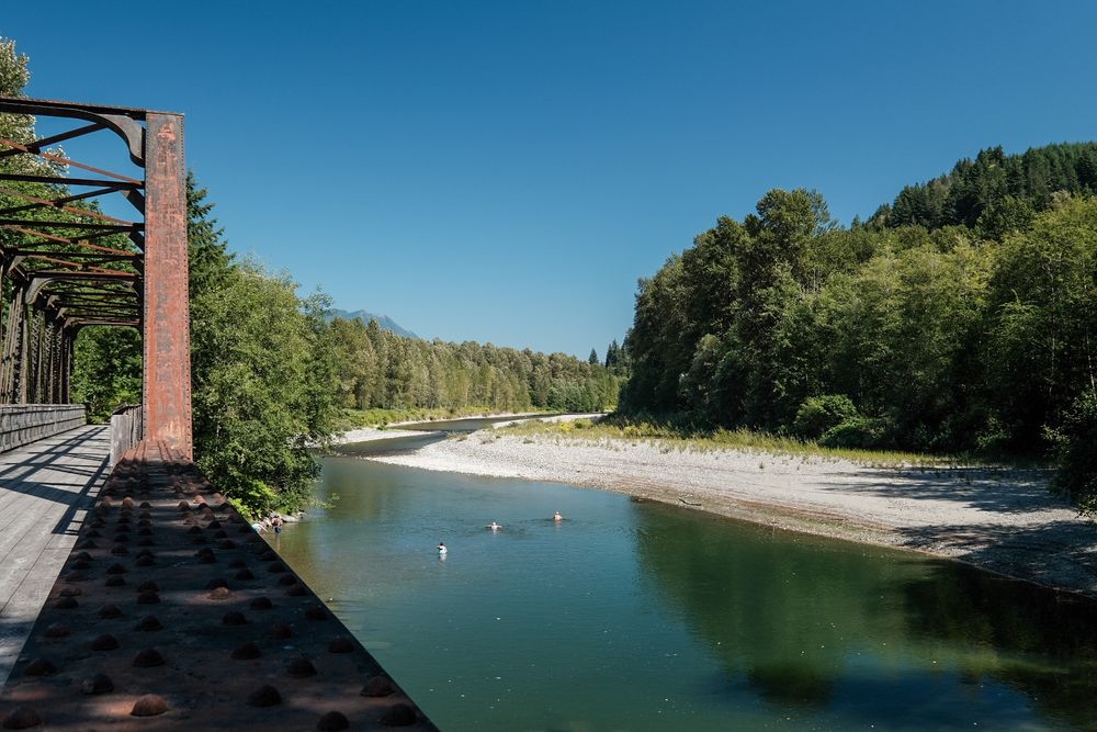 Looking across an old railroad trestle that crosses the North Fork of the Stillaguamish River. This is part of the multiuse Whitehorse Trail. 3 humans are swimming in the river which is lined by trees under a cloudless bluesky.