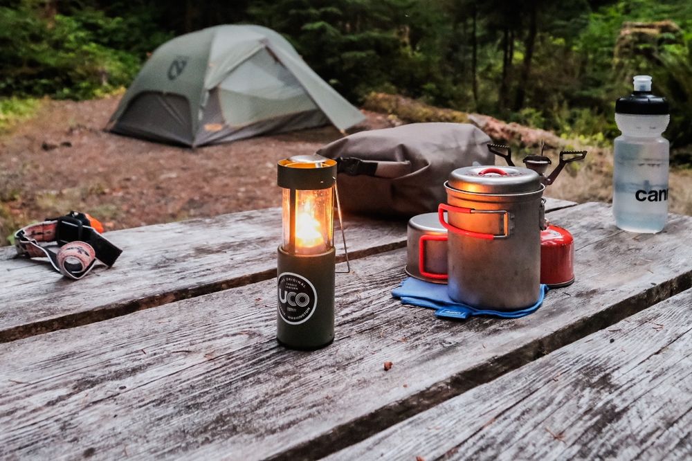 A small candle lantern illuminates a cook kit on a rustic camp ground picnic table. A tent is in the background 