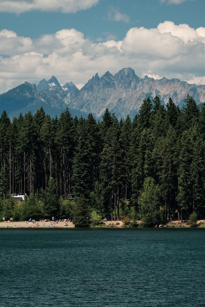 A view across the deep blue Easton Lake. A jagged mountain range in the Alpine Lakes Wilderness rises above ground a stand of trees along the lake. Puffy clouds cast shadows on the mountains.