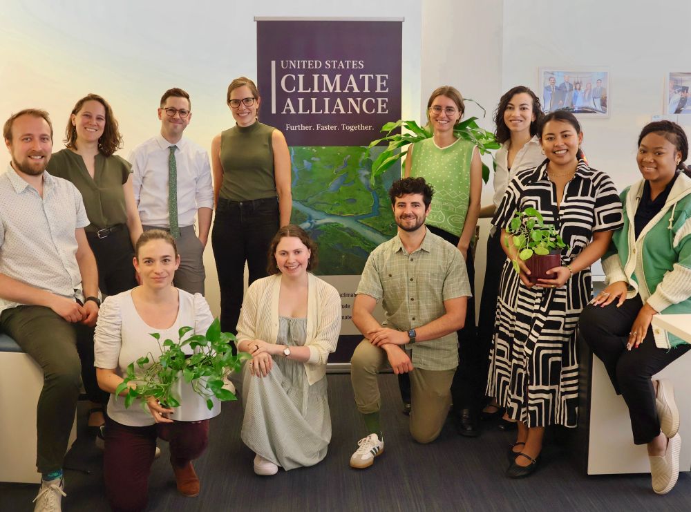Eleven U.S. Climate Alliance staff members wearing green for Earth Day, holding plants and standing in front of an Alliance banner. 