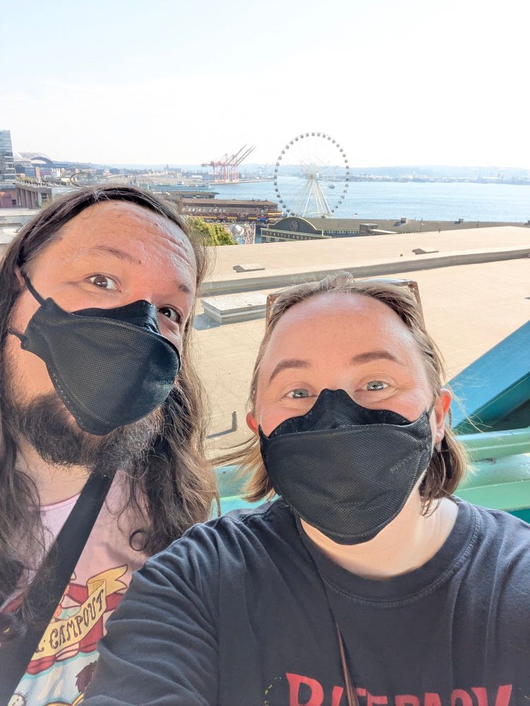 Me and Nate taking a selfie in front of the waterfront with the ferris wheel and Seattle Aquarium in the background. We're both wearing black KF94 masks and making wide-eyed faces.