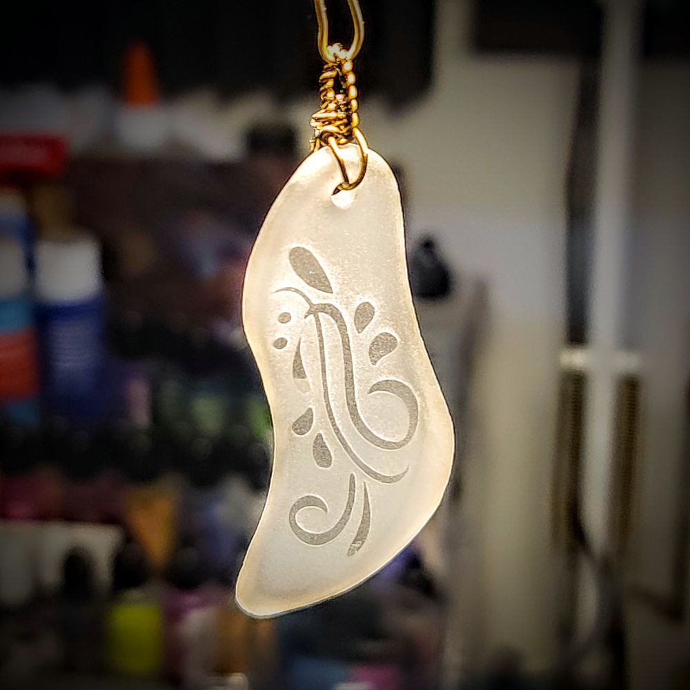 glass pendant made from a broken shard of booze bottle glass. vaguly l shaped and it has been frosted with some filigree swirls on its face left clear.