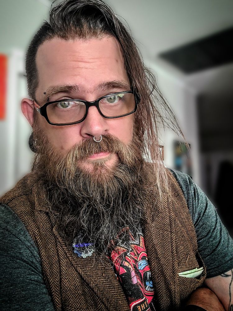 bearded man with side shaved hair and glasses wearing a grey shirt and brown vest