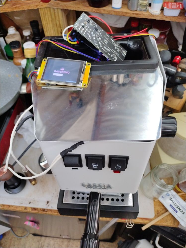 A White  Gaggia Classic Pro espresso machine with a computer board wired into the top. On the small screen it says "Gaggiuino."