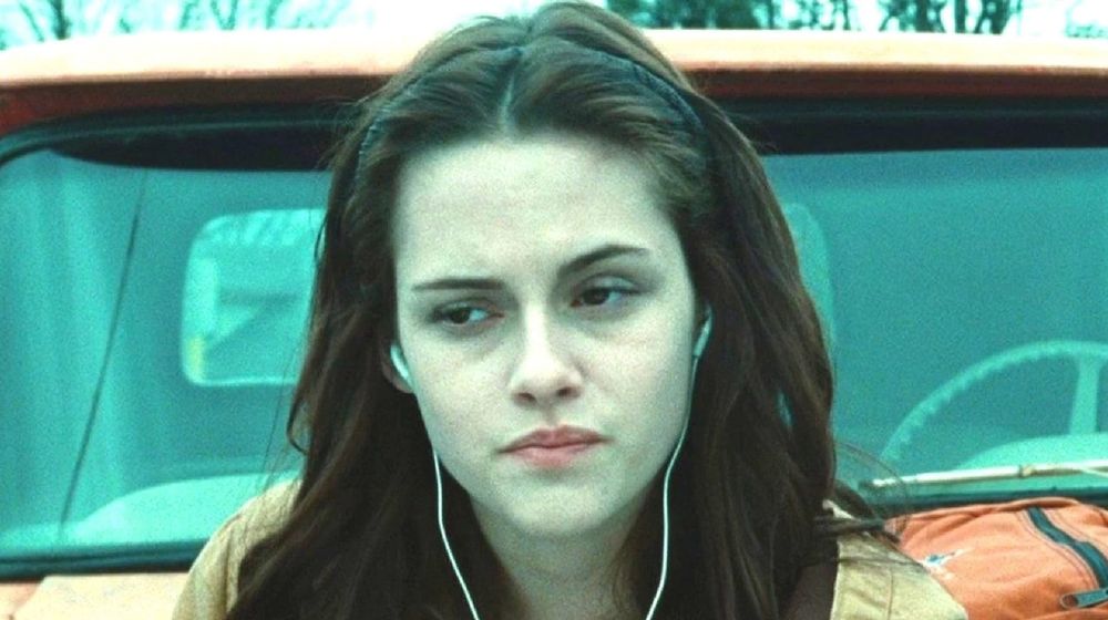 Kristen Stewart as Bella Swan in Twilight, standing in front of a car with headphones in her ears, looking off-screen like she is incredibly bored.