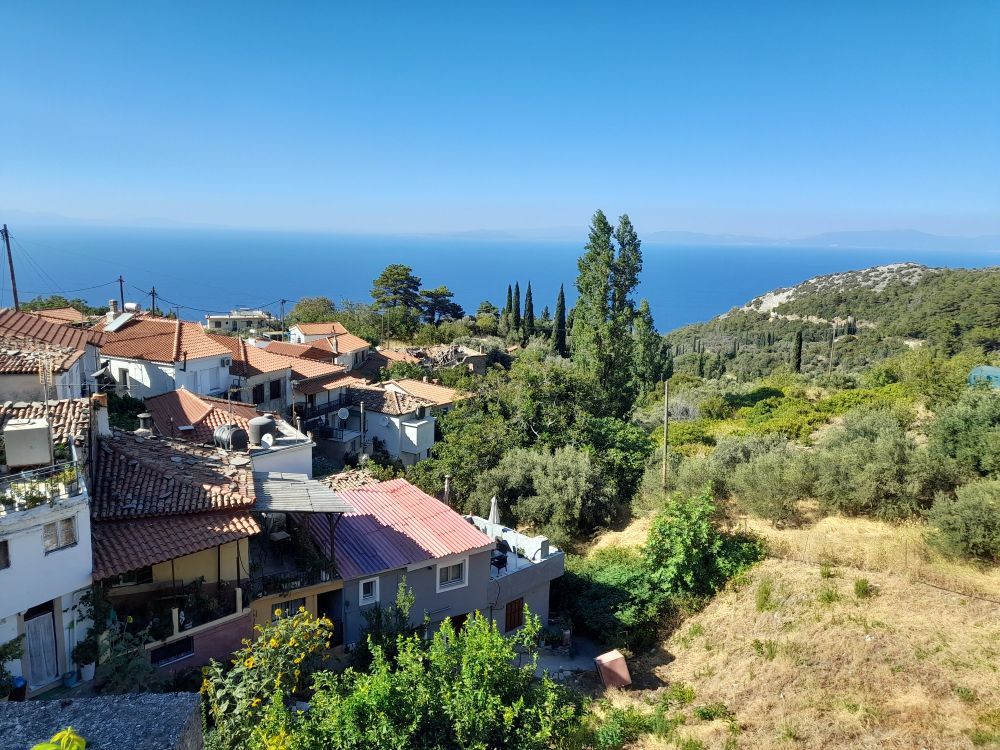 I'm Vordergrund ein griechisches Bergdorf und grüne Hügel, im Hintergrund das Meer