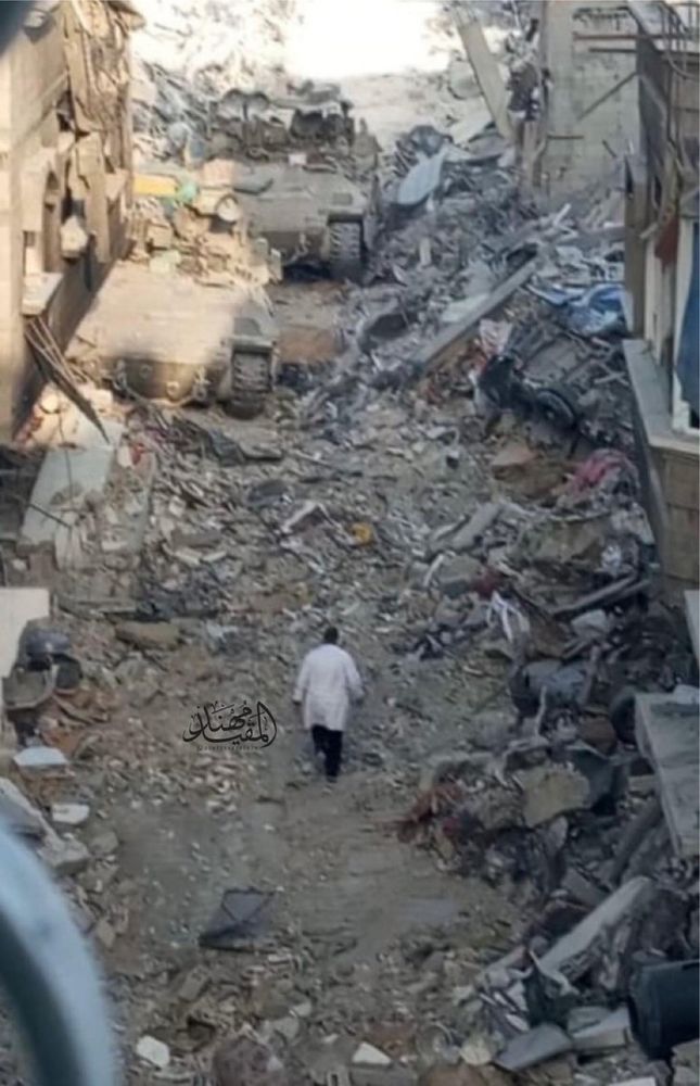 A man on a white lab coat walks away from the camera, surrounded by rubble. Two Israeli tanks look ahead. 