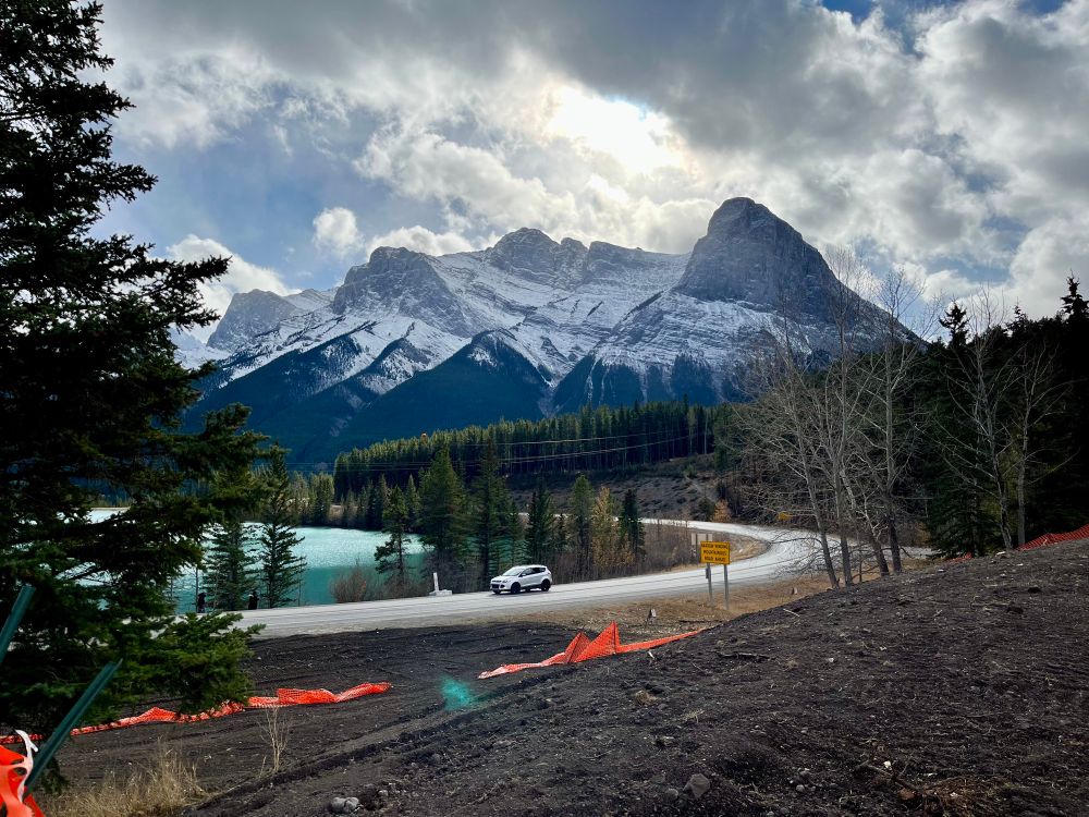 Light pours out of the clouds and halos a snow covered peak. A little blue lake lies at the foot encircled by a curve of road with a single white vehicle driving by.
