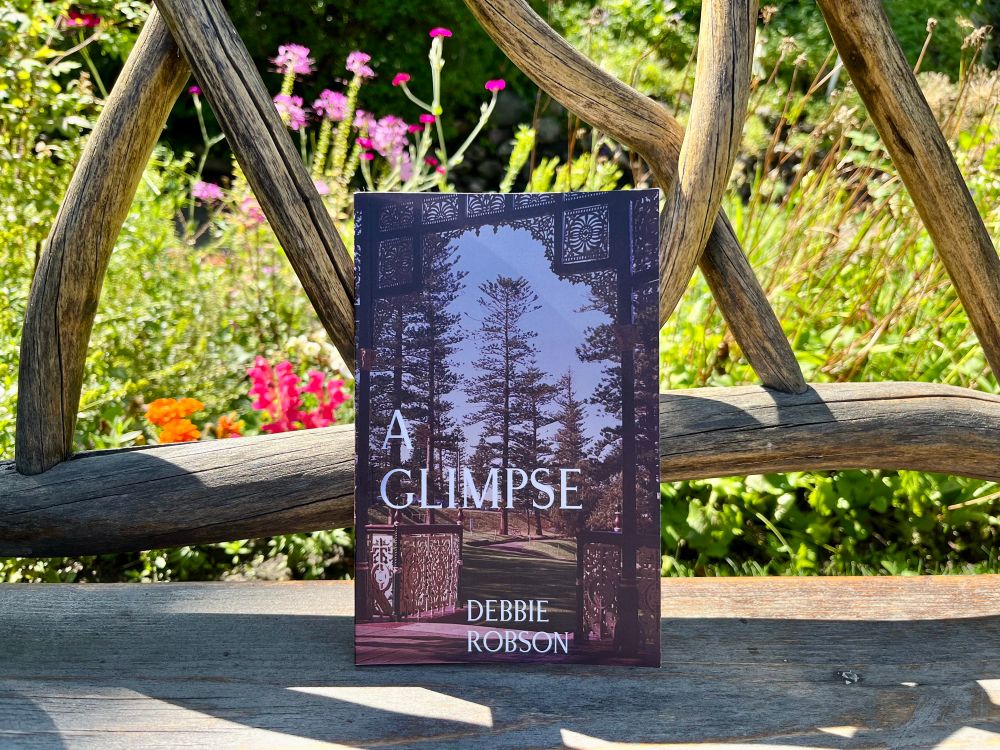 The chapbook “A Glimpse” by Debbie Robson sits against the back of an old wooden bench made of smooth twisty branches that form an ‘x’ pattern.  Behind the bench is a green garden with pink and orange flowers.