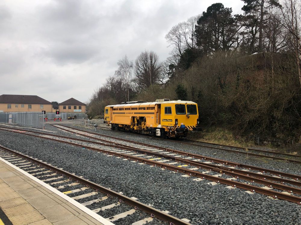 A mustard yellow tamper train parked in a train siding with bushes and trees behind it in the background. The two sidings in front of the train have been recently relaid.