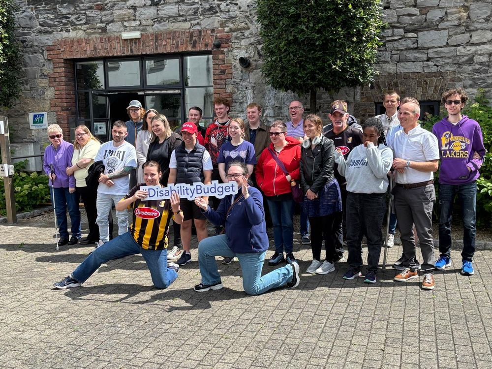 21 people in a group photo outside the DFI offices in Dublin standing behind two people kneeling down holding a sign saying "#DisabilityBudget"