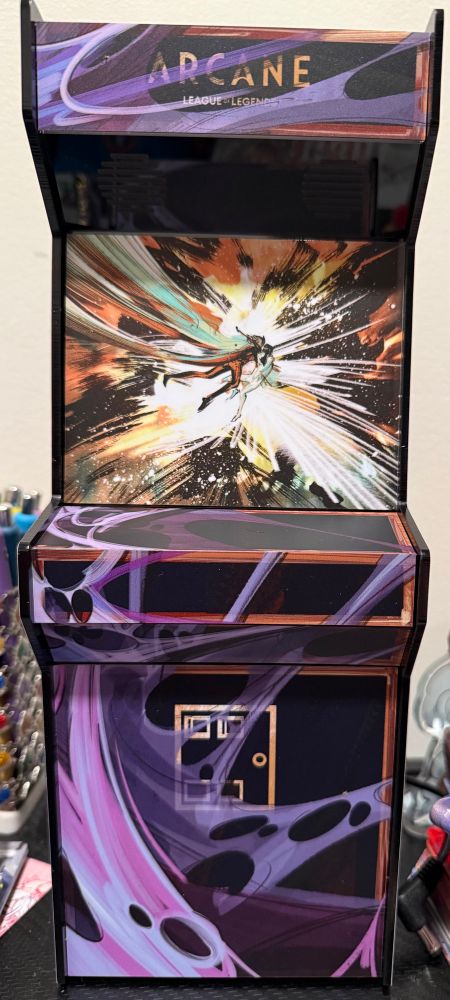 Front facing picture of the Hexcabinet from the Arcane exhibit for Gallery nucleus. It is a Mini Arcade cabinet featuring Jayce and Viktor embracing in the arcane on the screen with purple hexcore corruption coming from the left in a web like pattern. There’s an Arcane logo at the top that’s being covered by the corruption.