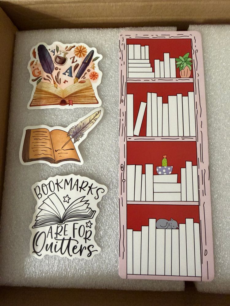 A bookmark shaped like a bookshelf with art of books, plants and a sleeping gray kitty. Three stickers, one a book open with pens, inks letters and flowers. Another is an open book with writing in it an a quill. The third has an open book and the words ‘bookmarks are for quitters’ 