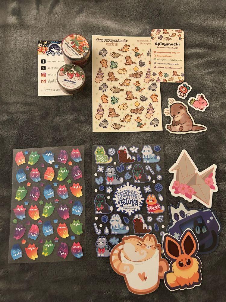 Merch haul image showing 3 artists’ worth of stickers and washi tape. Washi tape are strawberries and pokemon. Stickers are small animals, otters, cats, and more cats.
