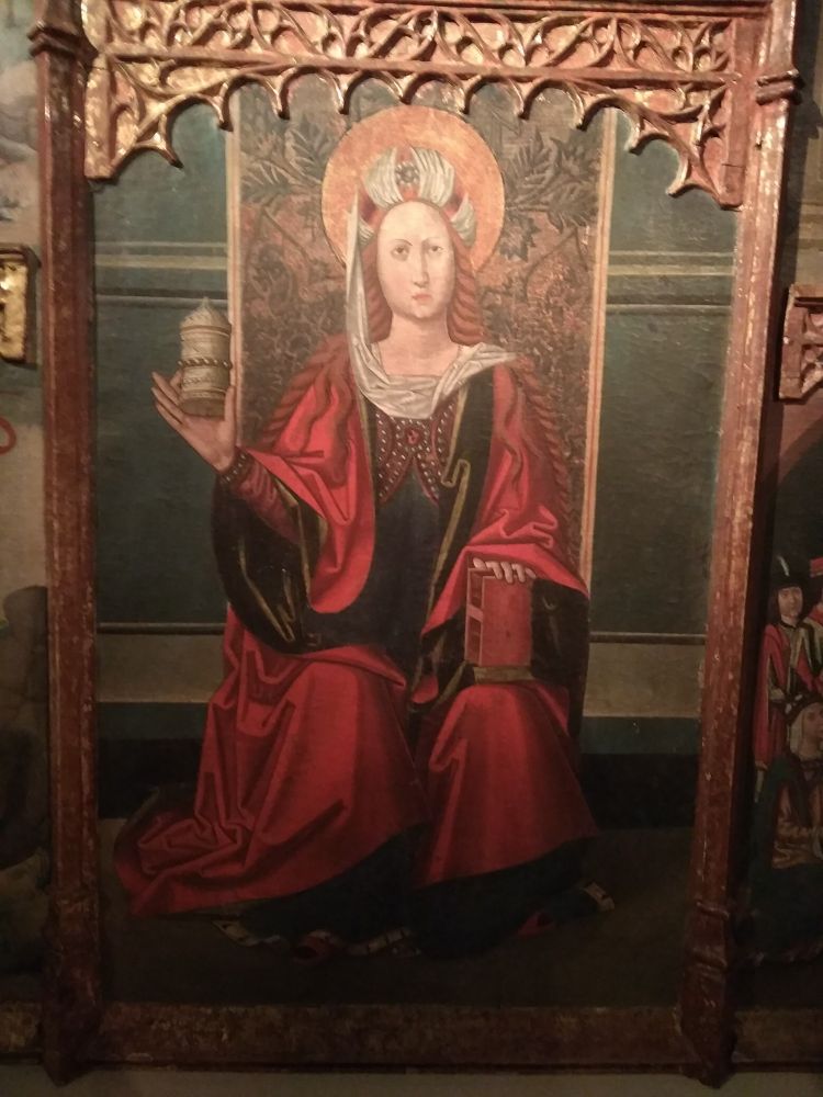 Centre-field of a fifteenth-century retable featuring the life of Mary Magdalen. The centre-field image depicts the Magdalen seated, dressed in a long red robe over a green kirtle. Her auburn hair flows in rivulets down her shoulders. She wears a white turban-like headgear with a jewel in front. In her right hand she is holding up the alabaster jar that is her main attribute - held between thumb and index finger - and her left hand is pressing a book against her left knee. The book has a red cover and the cover is kept together with a clasp. She looks angrily into the air.