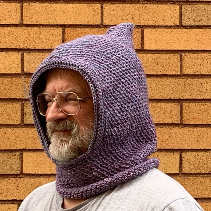 Head shot of me, white male w/white beard wearing grey/purple hooded cowl, facing 2/3rds camera left, in front of yellow brick wall