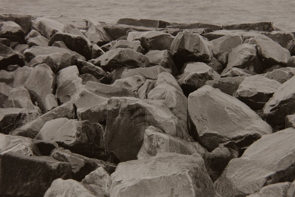 A black and white picture of the break rocks laying in heaps on the edge of Lake Erie. The water is just visible in the back. 