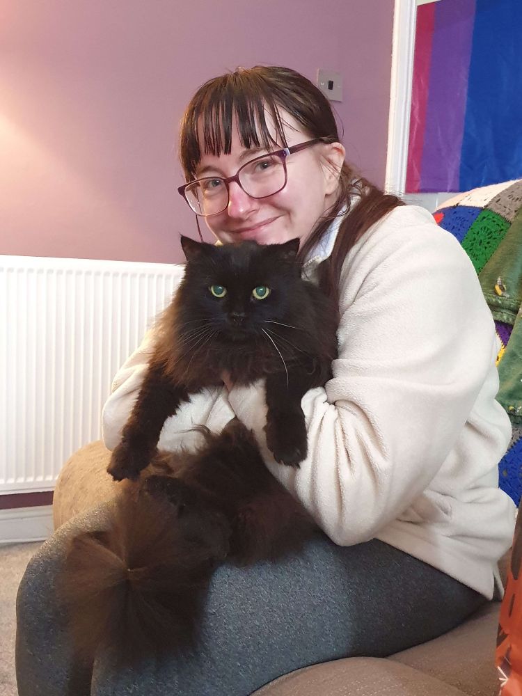 A picture of me, a white femme presenting person with long dark hair and glasses, cuddling Charlie, a long-haired black cat, on a brown couch. I am smiling, and Charlie is looking directly into the camera. Behind us is a Bi pride flag over a door.