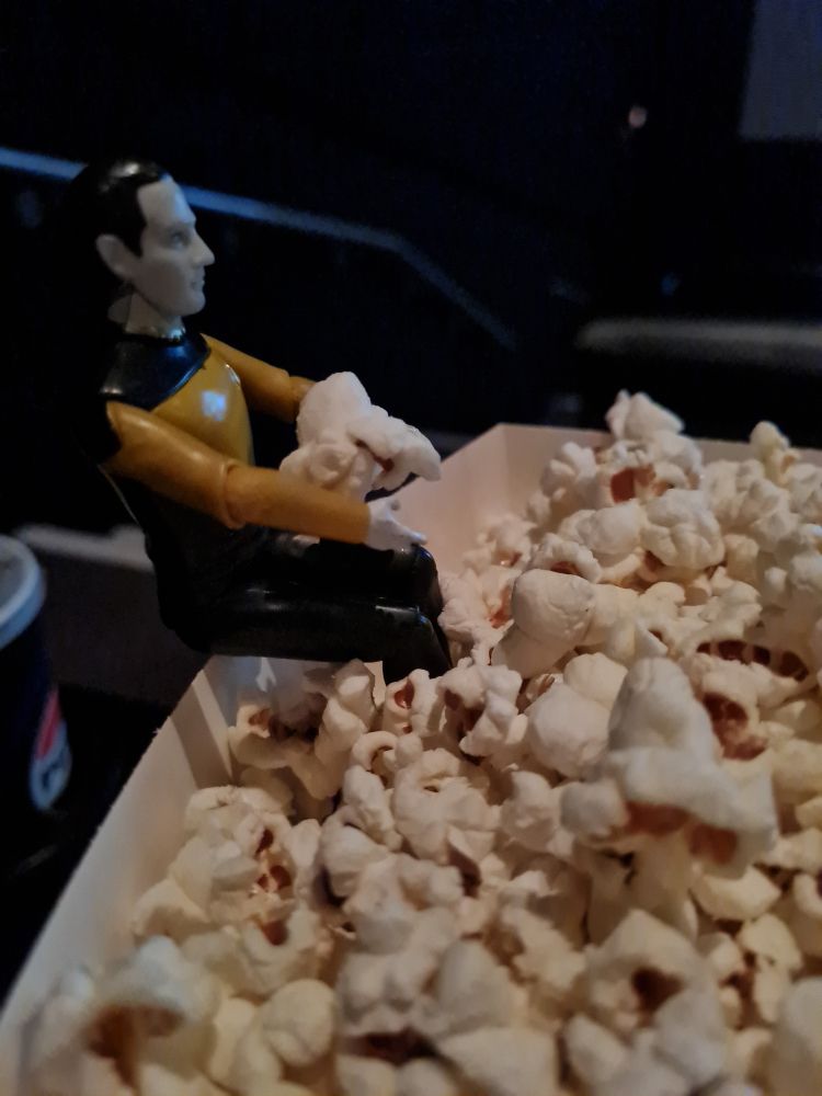 A little figurine of Data from Star Trek: TNG, is sat on the edge of a tub of popcorn in a cinema. He is holding one piece of popcorn in his arms.