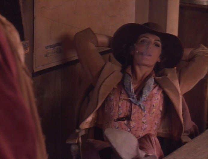 Deanna Troi from ST:TNG, wearing a cowboy outift. She's reclining in a chair with her feet up on a desk, while smoking a cigar.
