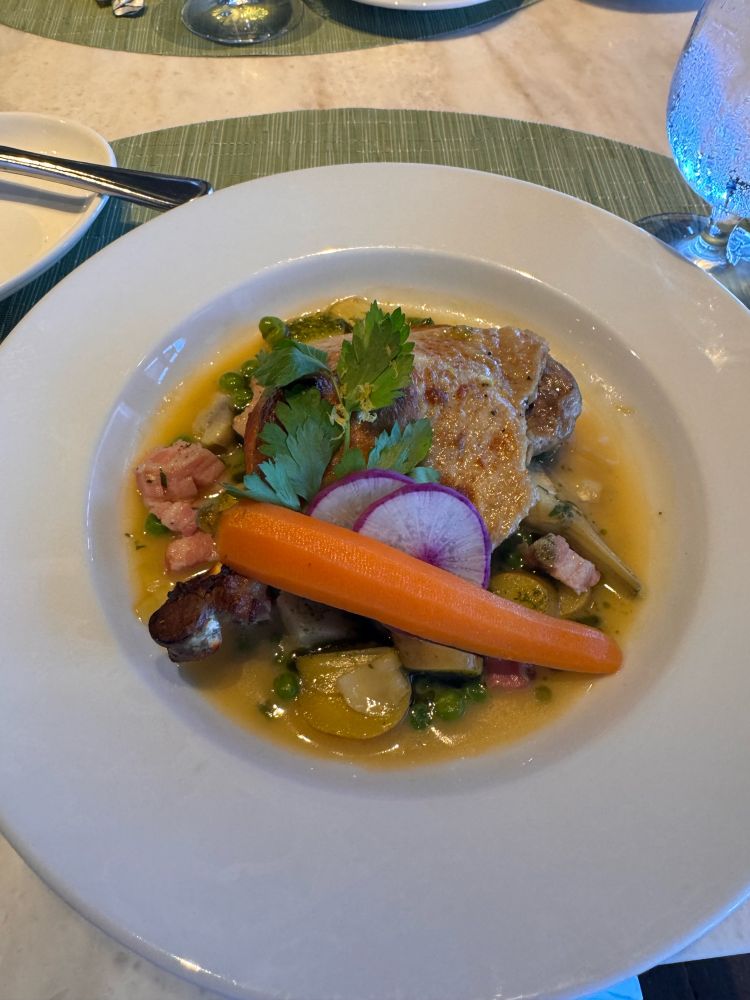 a large, white, ceramic plate holds a yellow broth with pieces of ham, potato, peas, artichokes and parsley. on the bed of veggies and broth there is a roasted duck leg, adorned with slices of radish, parsley and one peeled carrot. this image is of the duck leg dish at Mariposa Ala Moana.