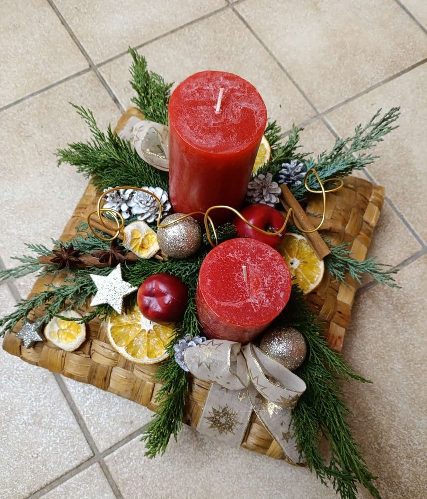 Viereckiges Adventsgesteck mit zwei roten Kerzen und viel Dekoration: Schleifen, Orangenscheiben, Sternen und Kugeln. 