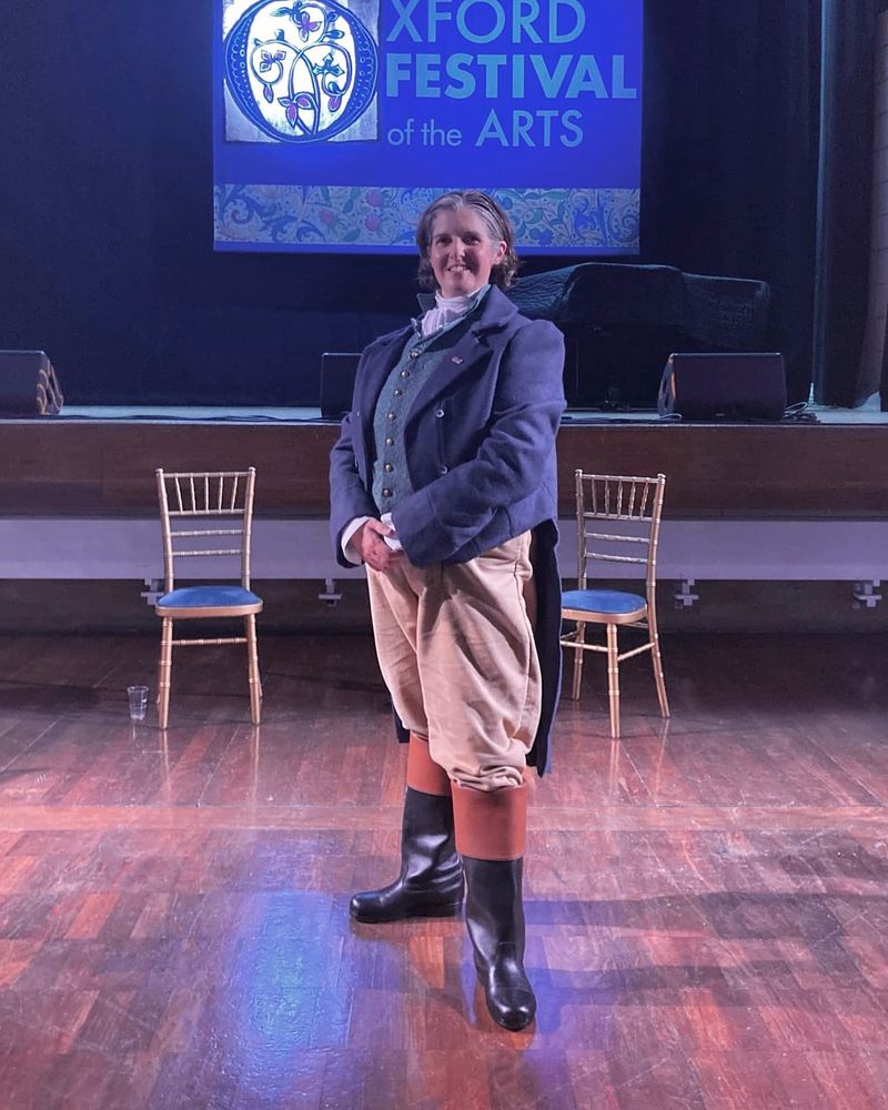 Erika Sanderson as Mr Knightley. She is dressed as a Regency gentleman, with a dark blue tailcoat, green waistcoat, cream trousers and black riding boots.