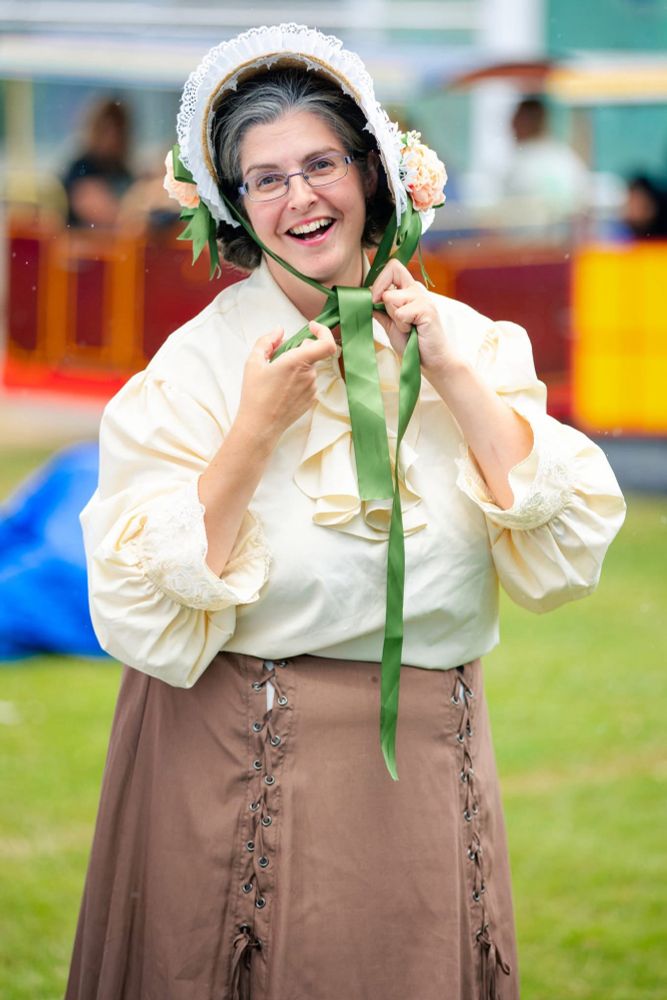 Erika Sanderson as Gertrude Jekyll. She wears a long brown skirt and cream Edwardian style blouse. She is tying the ribbon of a floral bonnet under her chin and smiling.