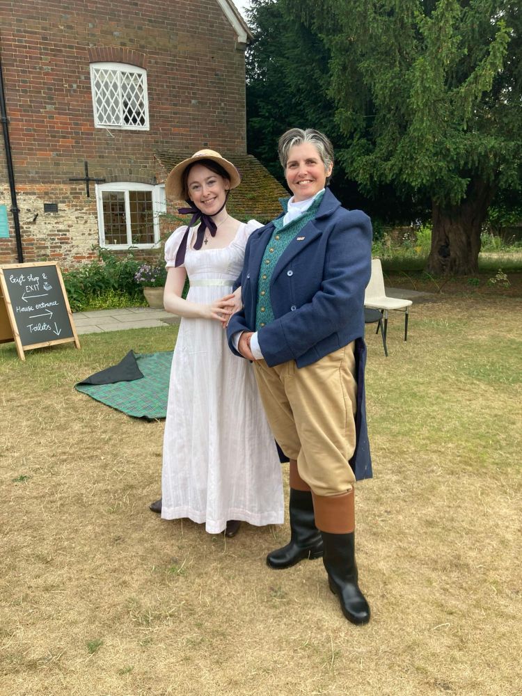 Helen Davidge as Emma. She wears a pink Regency dress, and straw bonnet with a purple ribbon.
Erika Sanderson as Knightley. She is wearing a blue tailcoat, with a white shirt and cravat, a green waistcoat, beige trousers, and black and brown cuffed riding boots.
Helen and Erika stand together in the garden linking arms as a couple and smiling. Behind them is the brick building of Jane Austen’s house, with a lattice window.