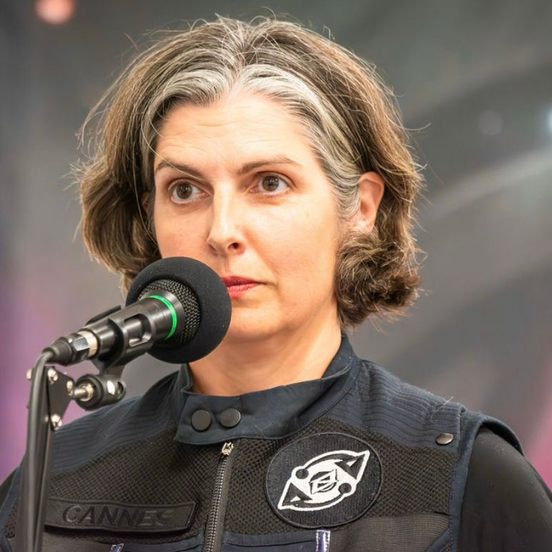 Erika Sanderson as Officer Hannah Cannes. She has short dark hair with a white streak and dark eyes. She stands in front of a microphone wearing a black military style vest.