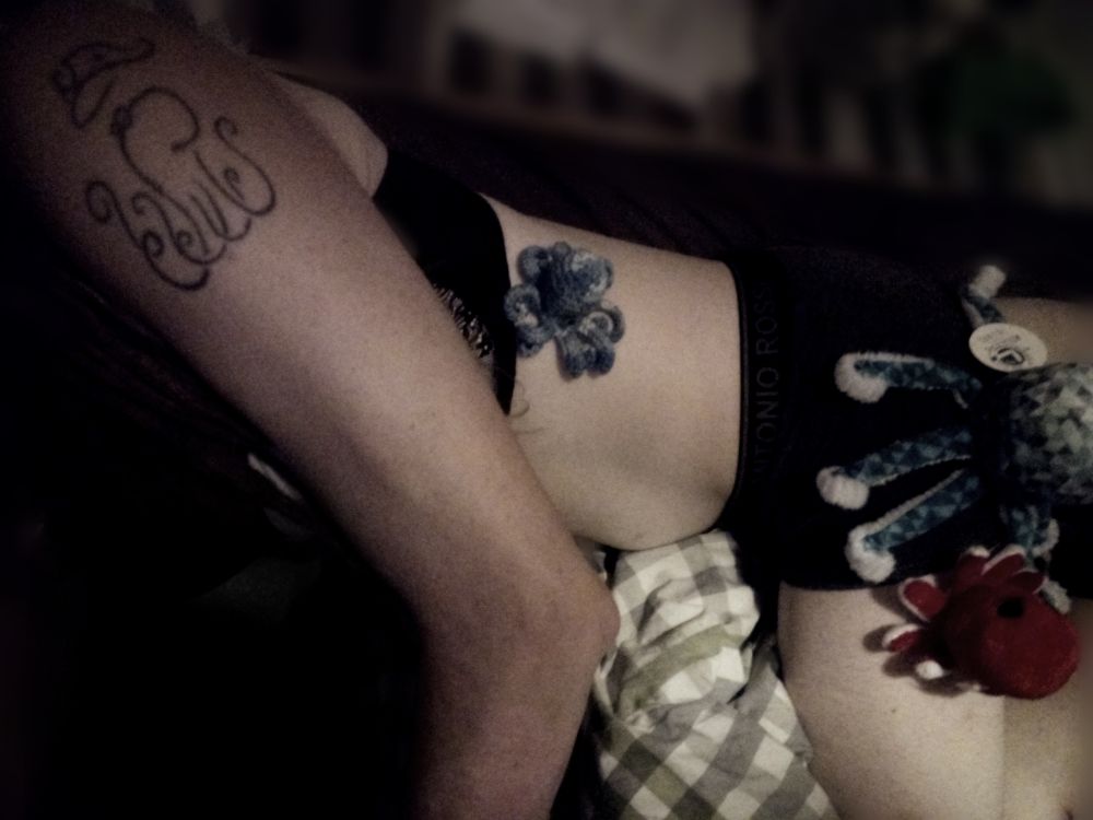 Foto einer Person, die nur in schwarzer Unterwäsche bekleidet auf der Seite auf einem Bett liegt. Auf dem Oberarm ist ein Tattoo von einer Krake mit Hut in einem minimalistischen, aber verschnörkelten Stil, seitlich auf dem Brustkorb sitzt eine kleine, blaue, gehäkelte Krake, auf dem Oberschenkel zwei weitere Plüschkraken, eine kleine, rote und eine etwas größere mit blauem Muster.