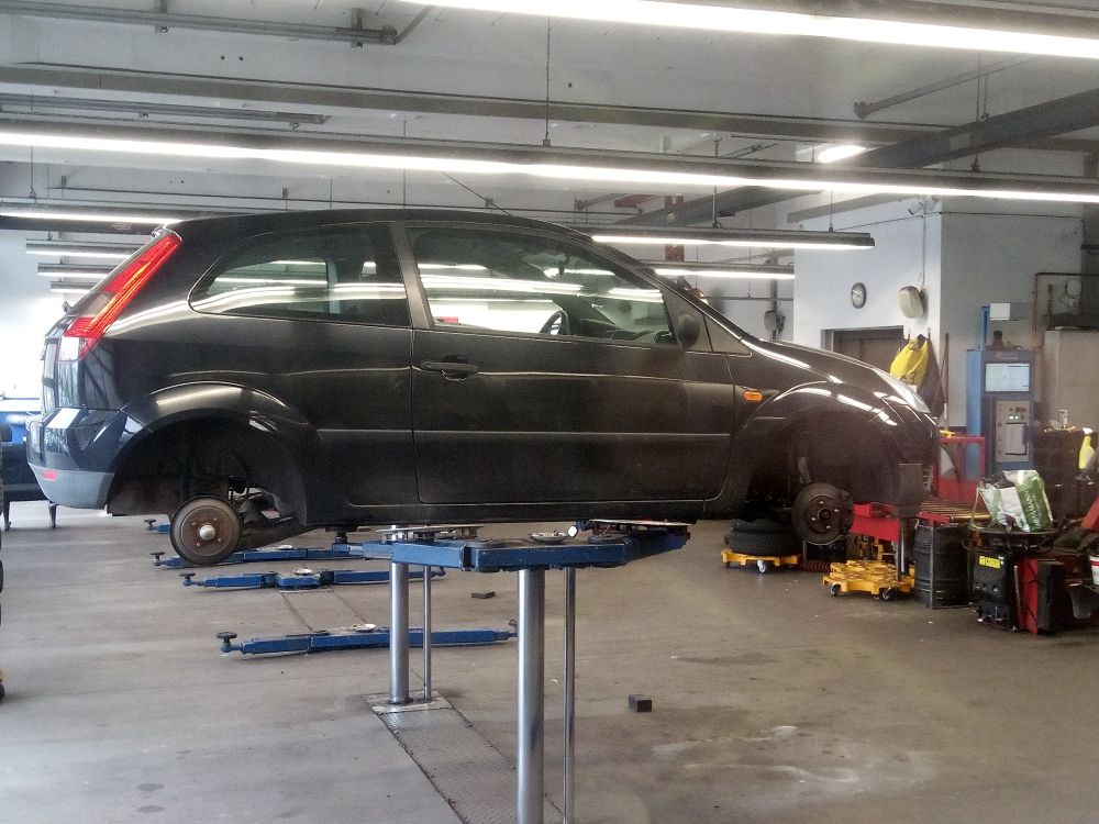 Photo of a black Ford Fiesta (older model) in a workshop. It's lifted up by some hydraulic pistons and has its wheels completely removed.
