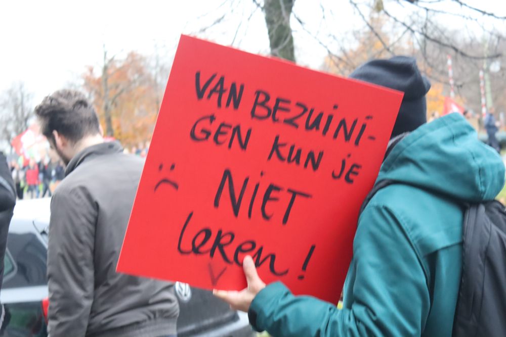 A bright red poster stating "van bezuiniggen kun je niet leren!" (you can't learn from budget cuts) with a sad smiley face.