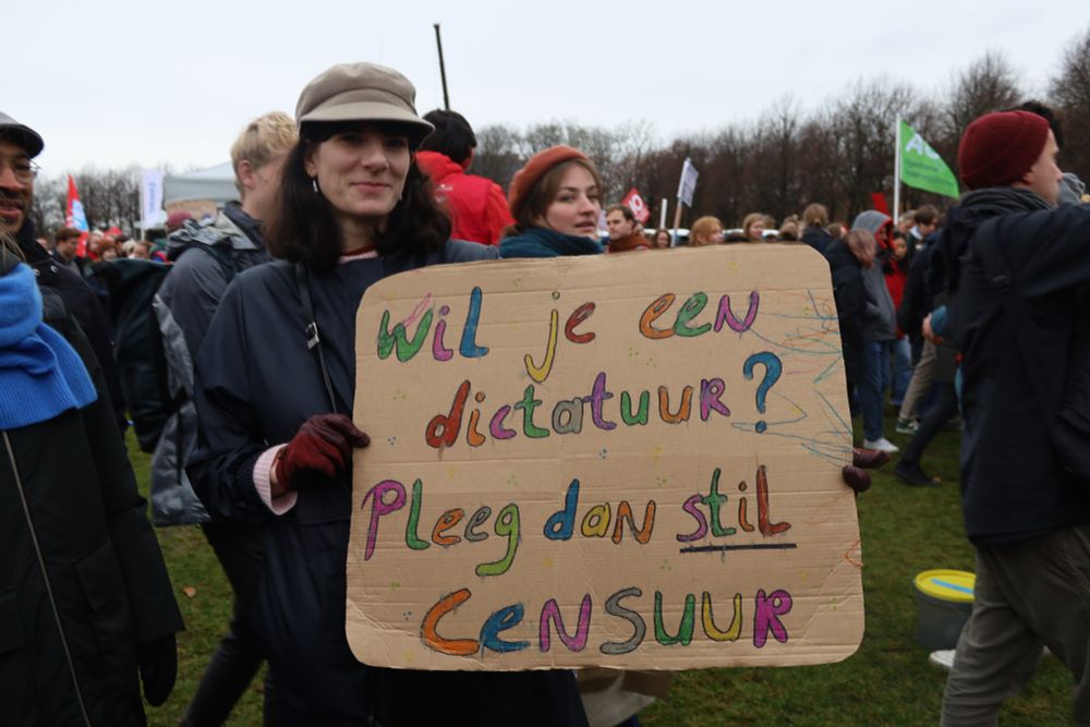 A woman, elegantly dressed and wearing a little smile, holding up a cardboard sign with colourful writing on it that says, in Dutch, "wil je een dictatuur? Pleeg dan stil censuur", which translates to "do you want a dictatorship? Then quietly exercise some censorship"