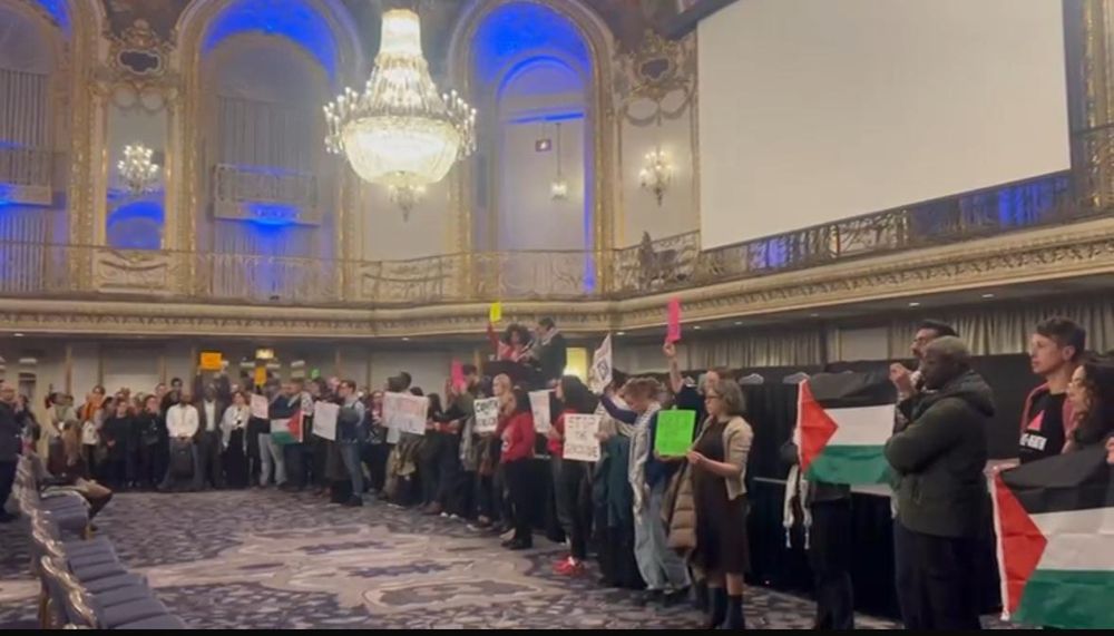Dozens of protesters in a ballroom holding signs condemning genocide, scholasticide, & censorship 