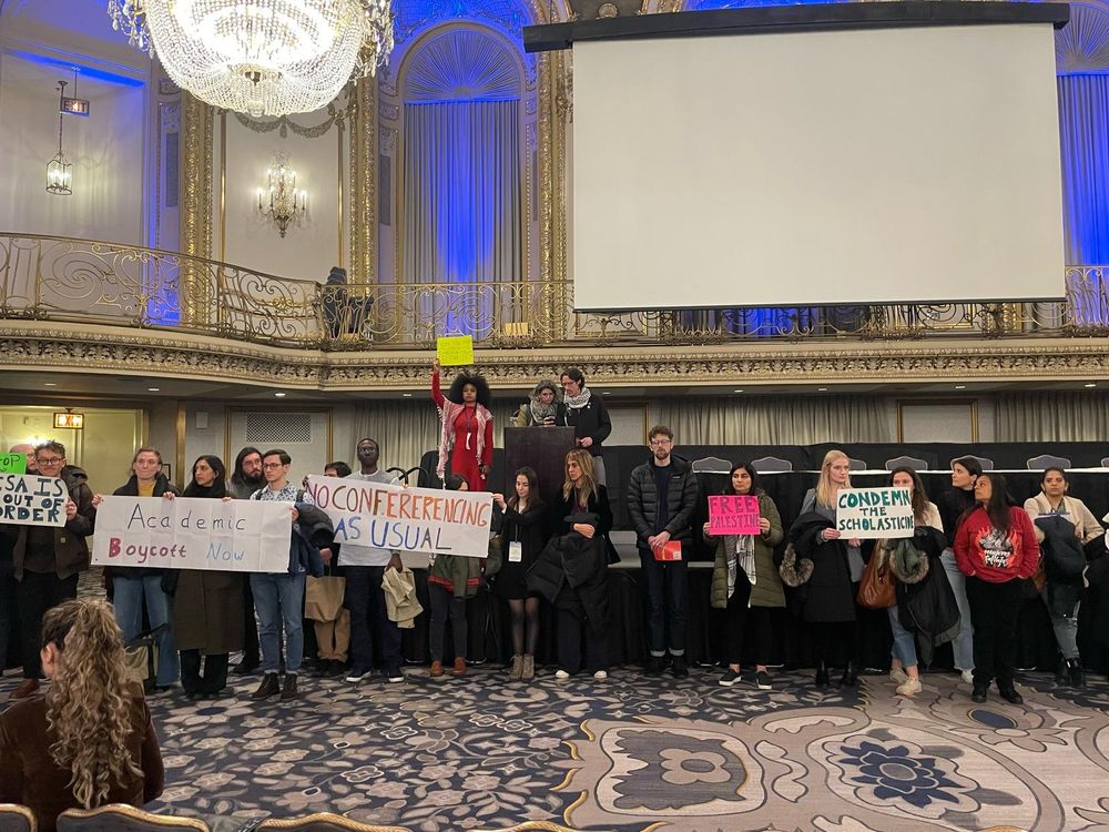 Image of protesters in a ballroom with protest signs condemning scholasticide, genocide and censorship