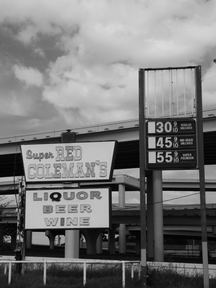Black and white image of the sign for an abandoned convenience store/gas station. One sign reads Super Red Coleman's Liquor Beer Wine. The sign next to is has incomplete prices for gasoline. Highway overpasses are visible in the background.