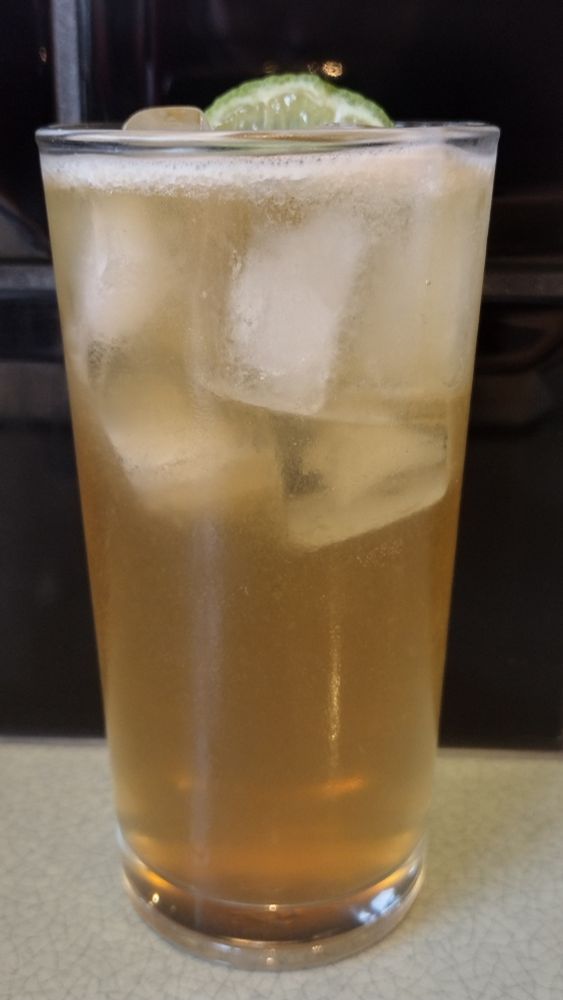 A sparkling golden Midsummer Daydream cocktail in a tall, ice-filled glass with a wheel of lime peeking out of the top.