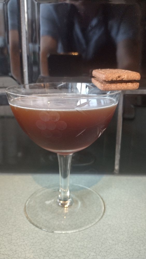 A dark brown Chocolate Biscuit cocktail in a coupe glass with a vine and grapes pattern, and a bourbon biscuit balanced on the rim as garnish.