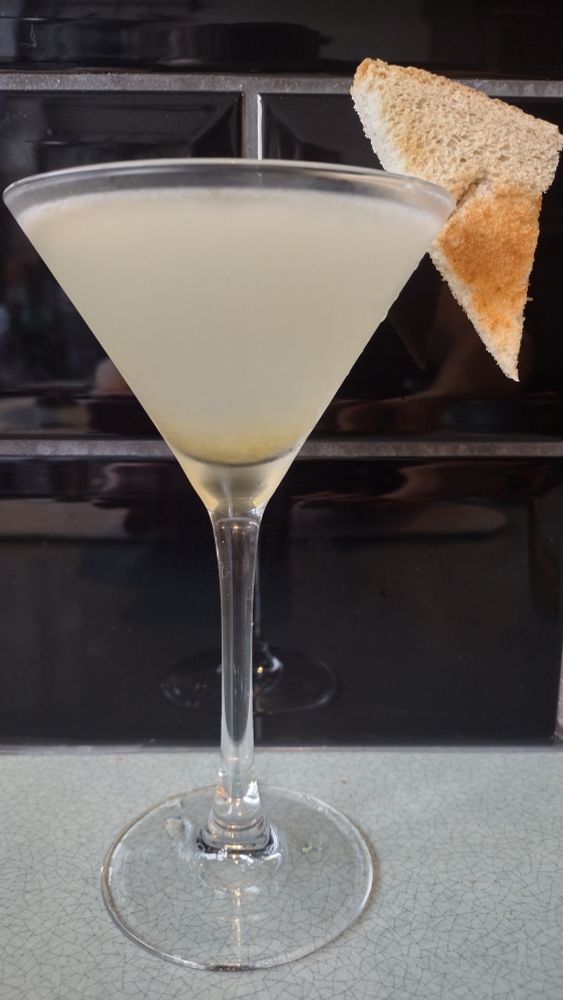 A yellowy Breakfast Martini in a cocktail glass, garnished with a triangle of toast on the rim.
