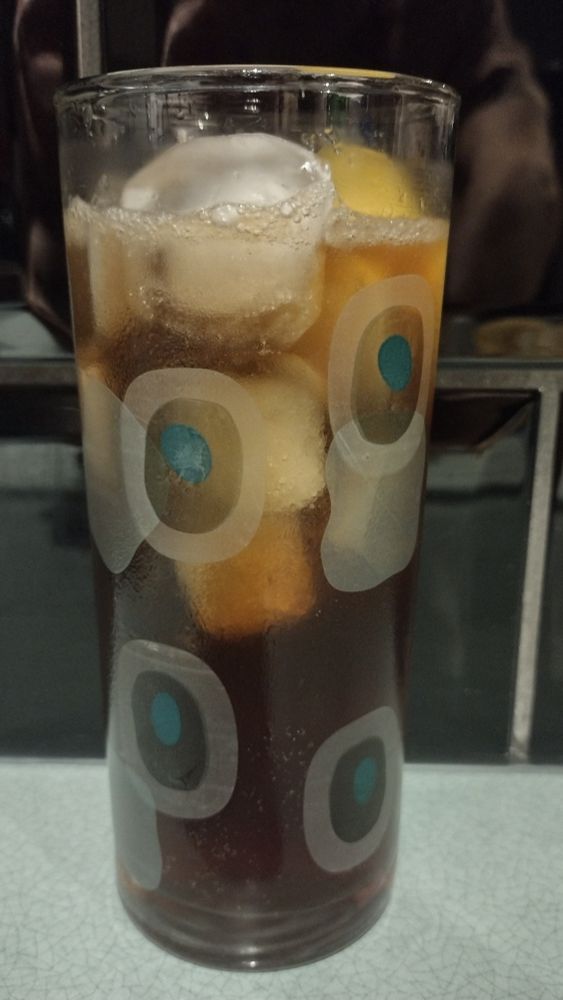 A dark London Iced Tea cocktail garnished with a lemon wedge in a tall ice-filled glass decorated with green shapes.