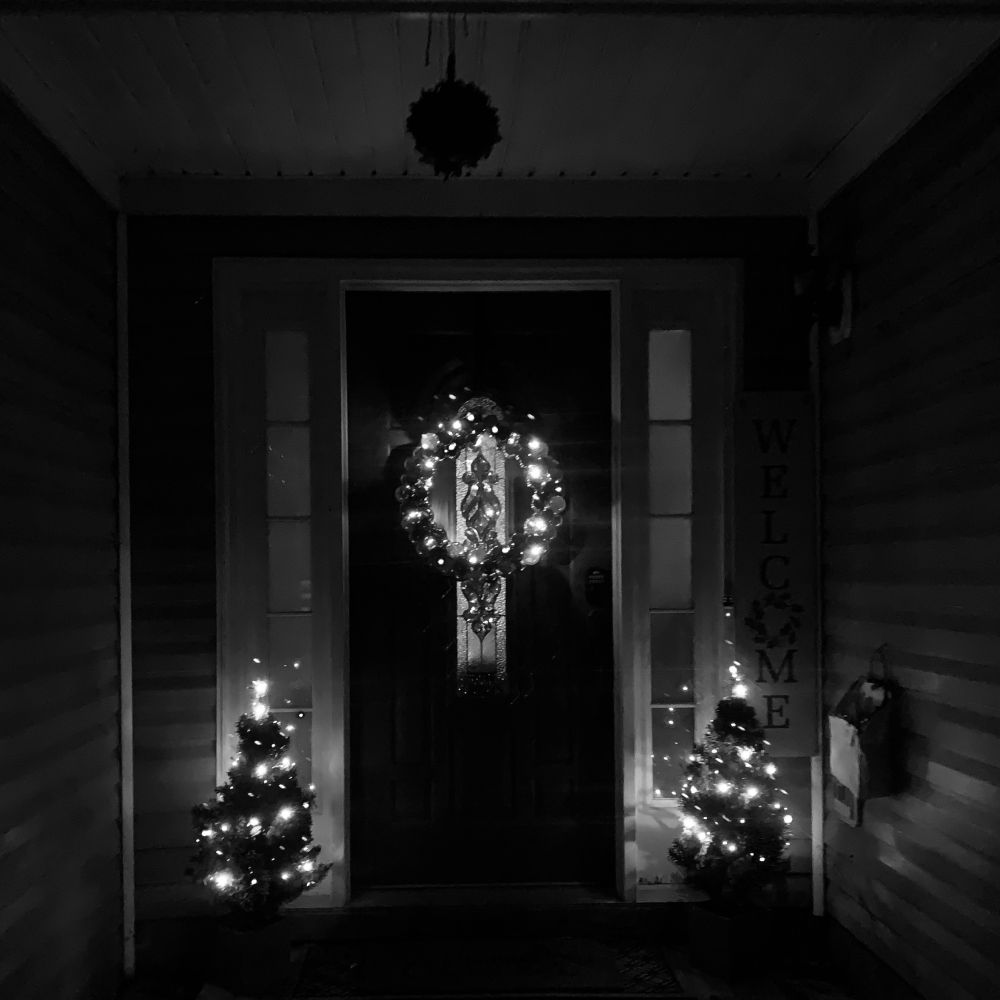 A black and white, darkened photo of a doorway with a lighted wreath and flanked by small lit Christmas trees
