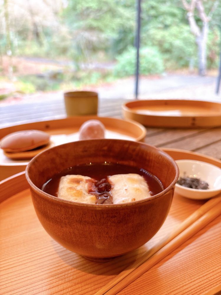 お椀に入ったおしるこ。焼いたお餅が二個入っている。奥に塩昆布の小皿やどら焼きがある。背景は緑の茂る庭