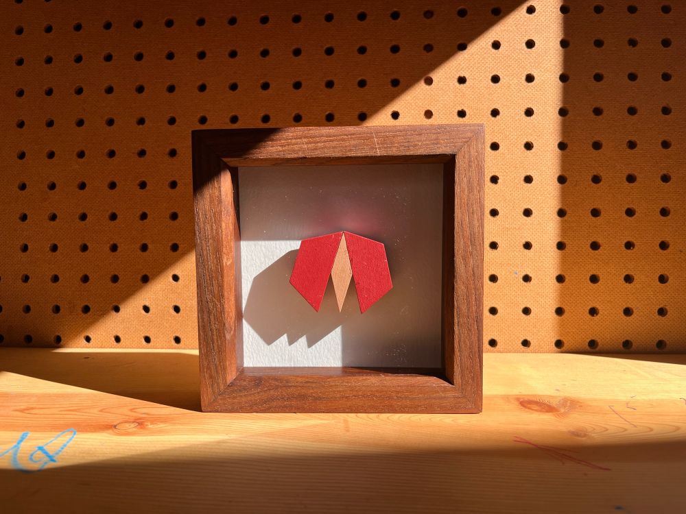 A bug made out of a tan rhombus and two red trapezoids. Framed in a 6 by 6 inch shadow box.