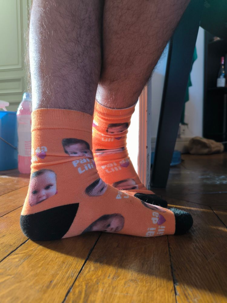 Foto dos pés do meu marido com o par de meias que eu mandei fazer. As meias são laranjas e tem fotos do rosto da nossa filha e o escrito "Pai da Lili 💜" repetidos em vários lugares.