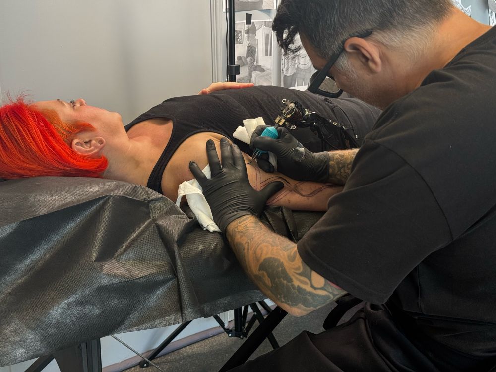 A woman with orange hair laying on a tattoo table being tattooed by a man wearing black gloves 